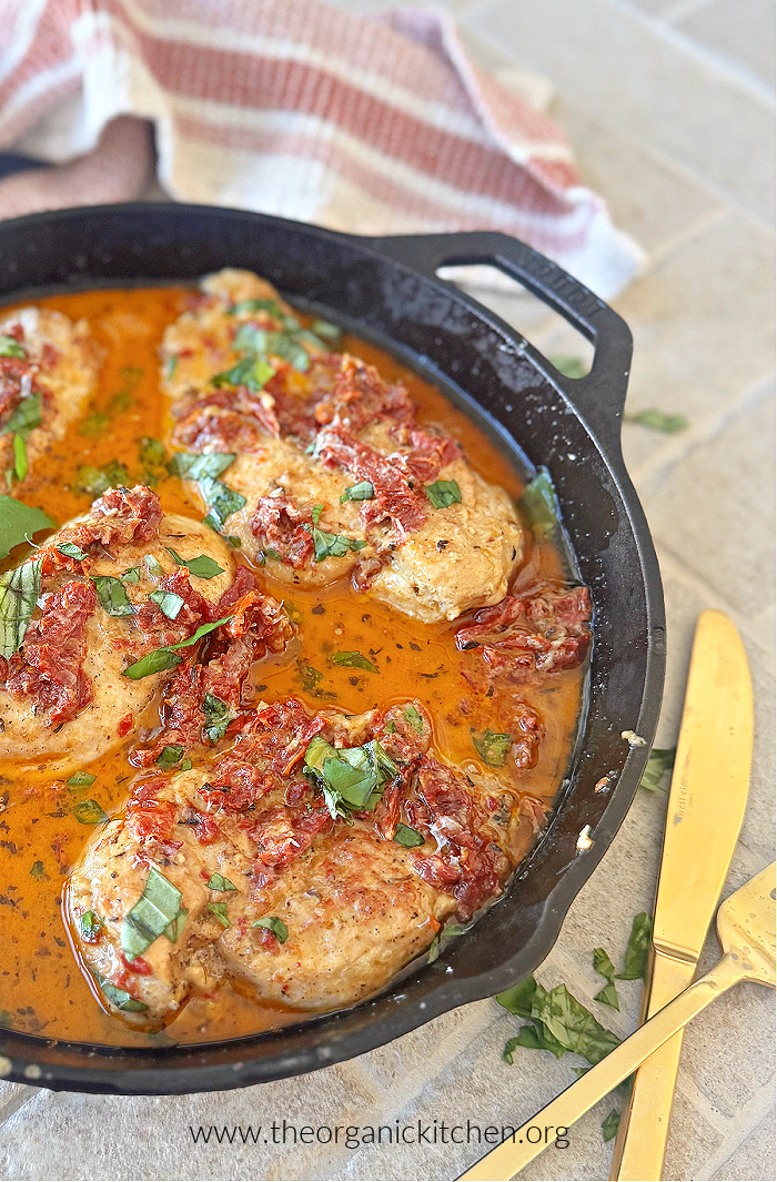 Marry Me Chicken garnished with basil in a black skillet with gold silverware and red and white dish towel in the background