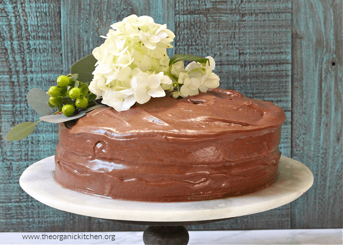 Classic Yellow Cake with Chocolate Frosting with white flowers and leaves on top, on a white cake plate with a blue background
