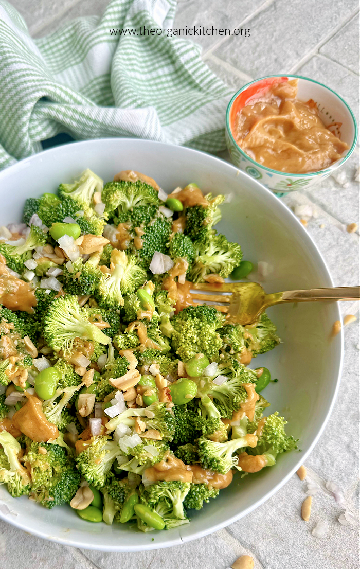 Twelve Delicious Vegetable Side Dishes Broccoli Salad with Peanut Sauce in white bowl with small bowl of peanut sauce and dish towel in background