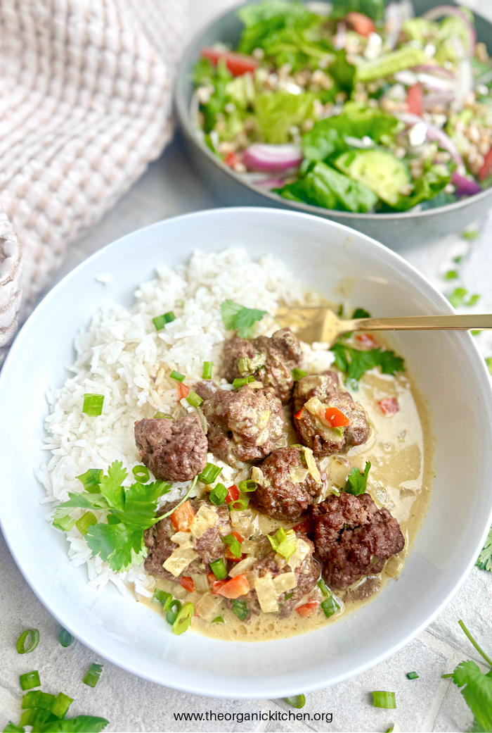 Easy Curried Meatballs and Rice topped with herbs in white bowl with gold fork, salad in the background