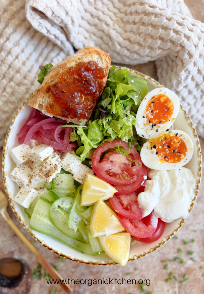 Healthy Mediterranean Breakfast Healthy Mediterranean Breakfast: greens, soft boiled eggs, cucumber, tomatoes, greens and a piece of toast in a bowl with dish towel above