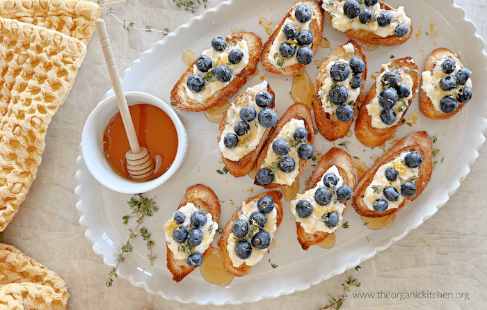 Crostini with Lemon Ricotta and Blueberries Crostini with Lemon Ricotta and Blueberries garnished with thyme on a white platter with a small bowl of hiney
