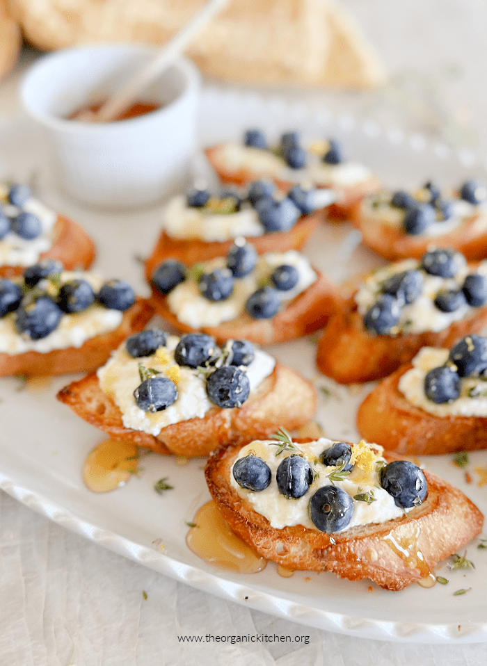 Crostini with Lemon Ricotta and Blueberries drizzled with honey and garnished with thyme on white plate