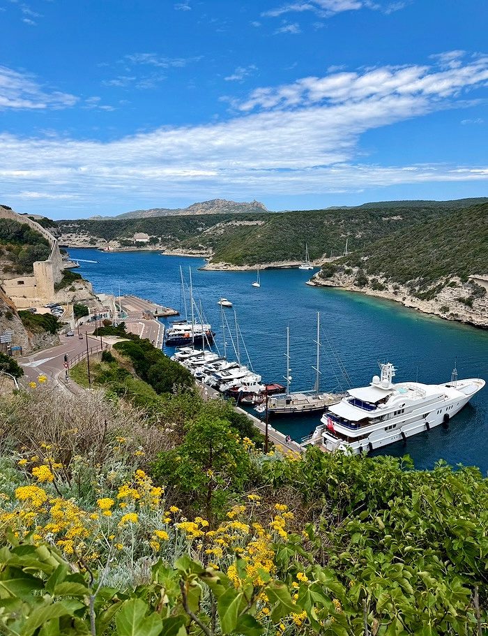 Boats in torquoise blue water surrounded by hillsides of yellow flowers and blue sky, Corsica France