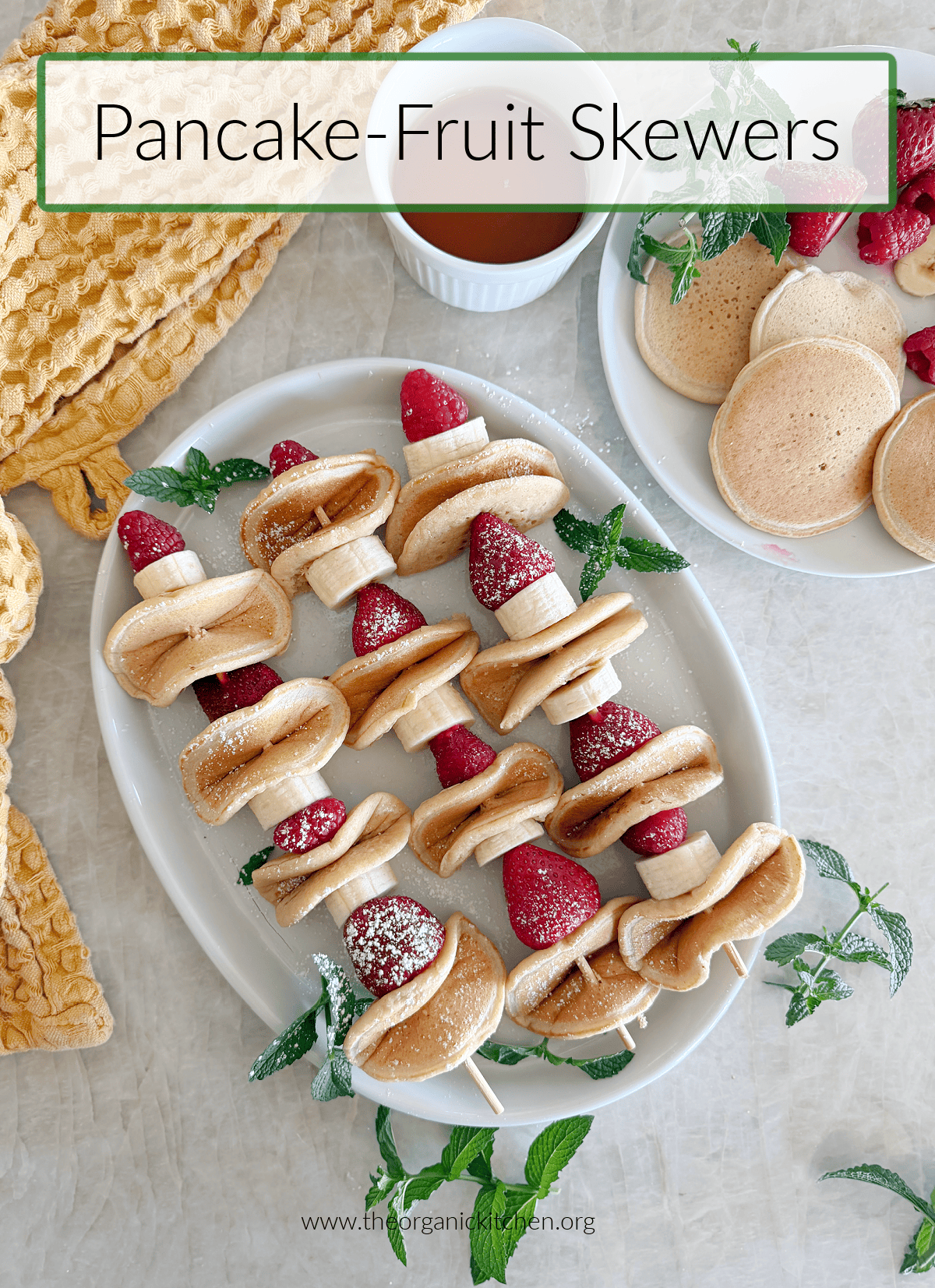 Pancake Fruit Skewers Three Pancake Fruit Skewers sprinkled with powdered sugar and garnished with mint on white platter with gold dish towel in the background
