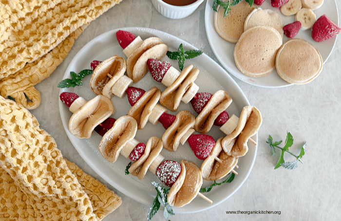 Pancake Fruit Skewers Three Pancake Fruit Skewers on a white platter surrounded by more berries, pancakes and a dish towel