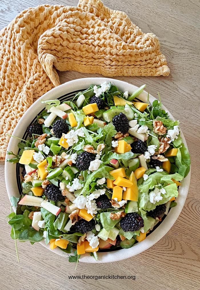 A white bowl overflowing with Blackberry Walnut Salad with Honey Vinaigrette, gold dish towel in the background