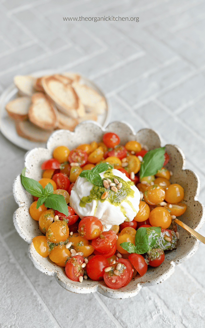 Tomato Burrata Salad with Basil Vinaigrette Tomato Burrata Salad with Basil Vinaigrette in a bowl with gold serving spoon and a plate of taosted bread in background